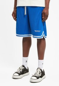 Pantalones cortos deportivos azules con acentos blancos, cinturilla elástica y logo. Combinados con calcetines blancos y zapatillas negras con punteras blancas.