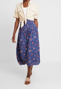 Lichtbeige shirt met korte mouwen, geknoopt in de taille, gecombineerd met een lange blauwe rok met een bloemenpatroon in rood en wit. Beige sandalen.