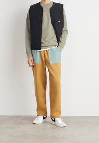 Schwarze gesteppte Weste über einem hellgrünen Sweatshirt, kombiniert mit orange-gelben Hosen mit hellblauen Akzenten und weißen Sneakers mit schwarzen Details.
