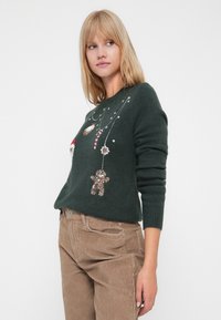 Vihreä neulepaita, jossa on koristeellisia elementtejä kuten lumiukko, karkkitanko ja glitterinen piparkakkuukko. Ribbaatut hihansuut ja pyöreä pääntie.