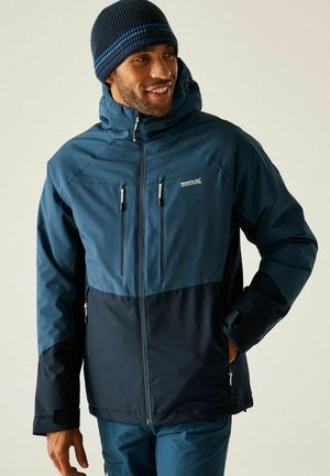 Hombre con chaqueta deportiva con cremallera azul marino y negra y gorro azul marino, sonriendo y mirando hacia un lado con una mano en el bolsillo.