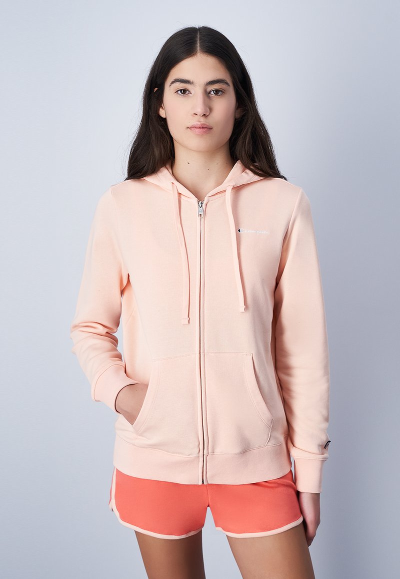 Sudadera con cremallera de melocotón claro, hecha de tejido suave, que presenta bolsillos frontales, cordones y un pequeño logo en el área izquierda del pecho.