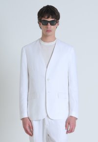 Blazer blanco de lino con un escote en V profundo, un solo botón y dos bolsillos frontales. Combinado con una camisa de color claro y gafas de sol rectangulares.