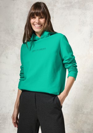 Groene hoodie met een zachte stof, voorzien van de tekst "CITY OF DREAMS" boven de zoom. Gecombineerd met donkere, relaxte broeken.