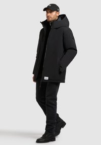 Svart zip-up jacka med en stor huva, kombinerad med en svart polotröja, mörka jeans och svarta stövlar. Har en logotag på botten.