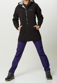 Schwarze Softshelljacke mit grauem Futter, langen Ärmeln und einem Reißverschluss vorne. Kombiniert mit lila Hosen und schwarzen Wanderschuhen.