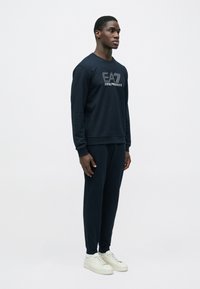 Tengerészkék pulóver logóval a mellkason, hozzá illő nadrággal és fehér sportcipővel kombinálva. Puha anyagból, kerek nyakkivágással és bordázott mandzsettákkal.