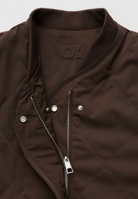 Bruine doorgestikte jas met geribbelde kraag, gedeeltelijk open zilveren rits en drukknopen aan de voorkant.