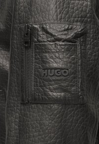 Mustast nahast jakk tekstuurse viimistlusega, millel on tõmblukuga tasku ja kummist logomärk "HUGO". Detailidena kontrastõmblused.