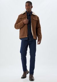 Brown fleece jacket with a cream collar, two chest pockets, worn over a denim shirt and dark jeans, complemented by brown boots.