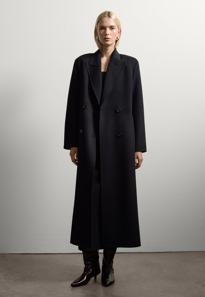 Cappotto lungo nero con design doppiopetto, ampi colli e tessuto testurizzato. Indossato sopra un indumento nero aderente e abbinato a stivali lucidi al ginocchio.