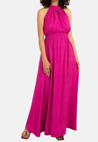 Lång halterneckklänning i stark rosa, med en samlad byst och en flytande kjol med strukturerade vertikala ränder. Platta sandaler synliga.