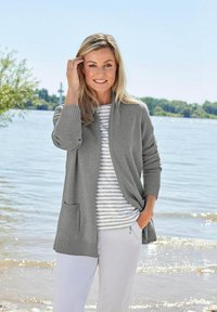 Grau gestrickte Strickjacke mit gerippten Bündchen und einer Vordertasche, über einem weiß-grau gestreiften Hemd getragen. Kombiniert mit hellen Hosen.