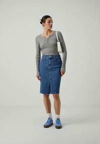 Mulher com uma camisa cinza de mangas longas ajustada, saia de ganga azul até ao joelho, sapatilhas azuis com meias brancas e uma bolsa branca de ombro, em pé de forma neutra.