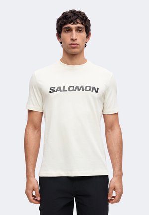 Jeune homme aux cheveux courts et bouclés, portant un t-shirt blanc avec le logo Salomon et un pantalon noir, debout devant un fond clair uni.