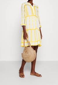 Mujer con un vestido de patrón amarillo y blanco sosteniendo un bolso redondo de mimbre, vistiendo sandalias de tiras color marrón, de pie frente a un fondo blanco liso.