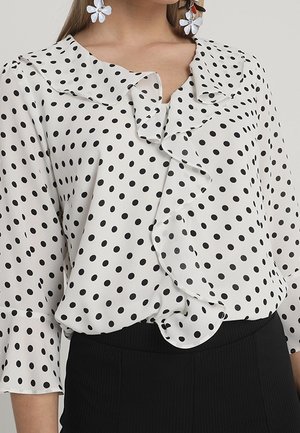 Blouse blanche à pois noirs, col et devant à volants, portée avec un pantalon noir et des boucles d'oreilles florales remarquables.