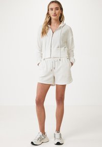 Felpa bianca con chiusura a zip e vita regolabile con coulisse, abbinata a pantaloni corti bianchi coordinati. Il tessuto sembra morbido con una finitura testurizzata.