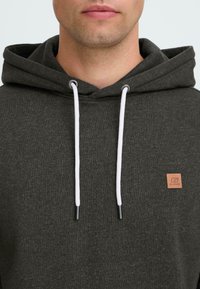 Man wearing dark gray hoodie with white drawstrings and small brown logo patch on the chest, visible from chin to upper torso.