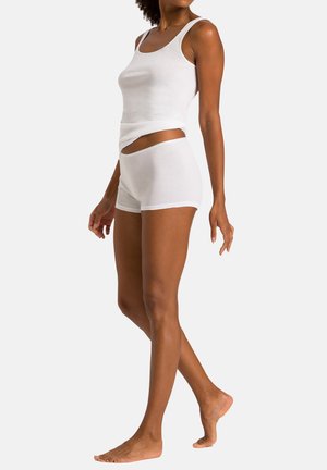 Kvinde iført hvid tætsiddende tanktop og matchende hvide shorts, stående barfodet mod en enkel lys baggrund.