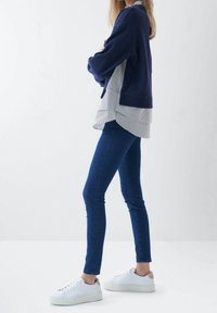 Felpa oversized blu navy sovrapposta a una camicia bianca a righe, abbinata a jeans skinny blu e sneakers bianche con accenti beige.