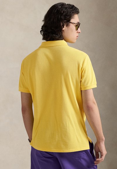 Κίτρινο πόλο πουκάμισο από υφασμάτινη υφή, με γιακά και κοντά μανίκια. Το πουκάμισο συνδυάζεται με μοβ σορτς.