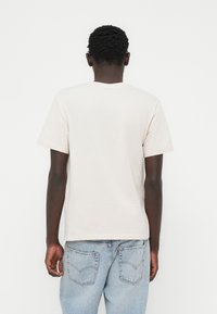 Κοντομάνικο ελαφρώς μπεζ T-shirt από βαμβάκι, με κλασική γραμμή και στρογγυλή λαιμόκοψη. Φορεμένο με ανοιχτό μπλε τζιν παντελόνι.