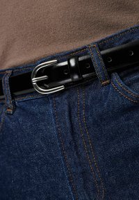 Melnas ādas josta ar sudraba sprādzi, valkāta virs tumši ziliem džinsiem ar kontrastainām šuvēm un vieglu tekstūru.