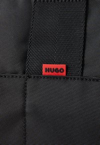 Juodas tekstūruotas audinys su raudonu stačiakampiu ženkleliu, ant kurio užrašyta "HUGO". Siuvinėtos detalės pagerina tvirtumą ir dizaino išskirtinumą.