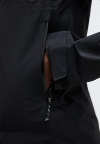 Ruka částečně zasunutá do kapsy černé bundy na zip, na paži viditelný rukáv s uzávěrem na suchý zip.