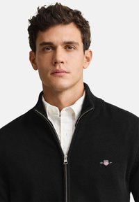 Μαύρο πλεκτό πουλόβερ με φερμουάρ και ριμπ λαιμόκοψη, με ασημί φερμουάρ και λογότυπο GANT στο αριστερό στήθος, φορεμένο πάνω από λευκό πουκάμισο.