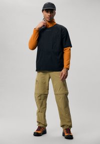 Moški v črni kapi, črnem kratkemu rokavu nad oranžno majico z dolgimi rokavi, bež cargo hlačah in oranžnih planinskih čevljih, stoji na sivi podlagi.