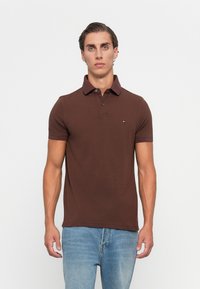 Mann som står med ansiktet fremover iført en kortermet brun polo-skjorte og lyseblå jeans foran en ensfarget bakgrunn.