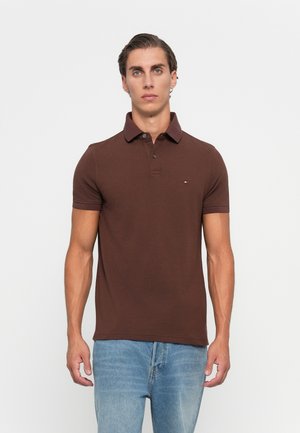 Moški, ki stoji spredaj, oblečen v kratkosleečno rjavo polo majico in svetlo modre kavbojke, proti enobarvni ozadju.