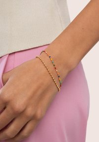 Bracciale d'oro con piccole perline e accenti colorati in rosso, verde e blu. Texture liscia, design delicato, indossato su un polso contro un tessuto rosa.
