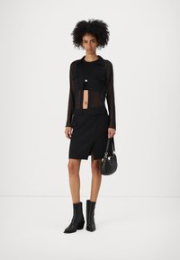 Top noir transparent à manches longues par-dessus un crop top, associé à une jupe noire ajustée avec une fente à l'avant. Bottines noires et petit sac.
