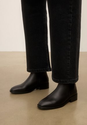 Bottines en cuir noir portées avec un jean noir à jambes droites sur un sol beige clair.