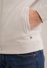 Hellbeige Kapuzenjacke mit Reißverschluss, ribbelten Bündchen und Saum, ausgestattet mit einer Seitentasche und einem kleinen Markenlogo nahe der unteren Naht.