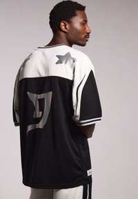 Diseño de camiseta en blanco y negro con mangas cortas, que cuenta con acentos de estrellas y un gran logotipo en la parte posterior. Fabricada con un tejido texturizado y transpirable.