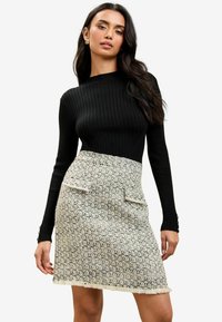 Čierny rebrovaný top s dlhým rukávom spárovaný s krémovou mini sukňou z tkaniny tweed, ktorá má textúrované vzory a dva predné vrecká s gombíkovými detailmi.