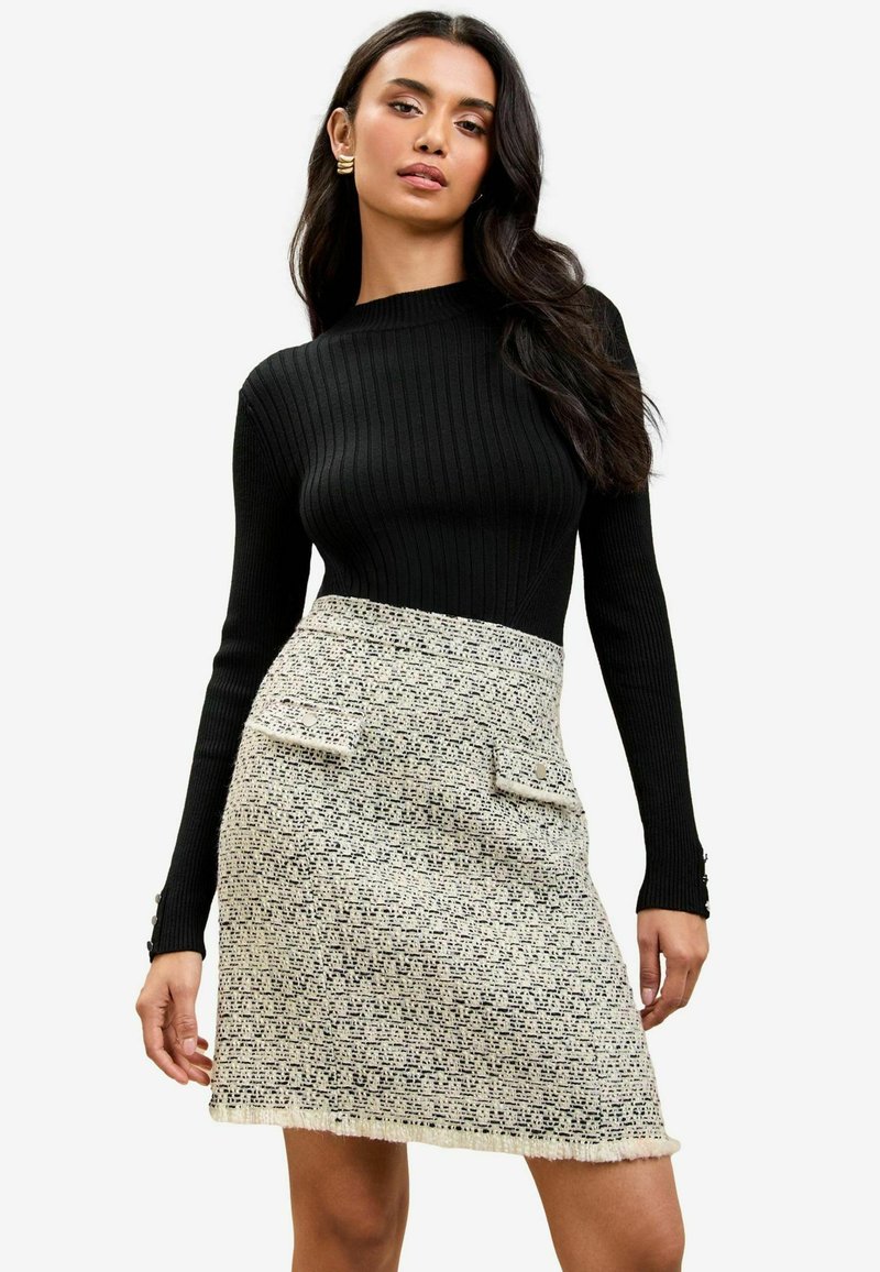 Čierny rebrovaný top s dlhým rukávom spárovaný s krémovou mini sukňou z tkaniny tweed, ktorá má textúrované vzory a dva predné vrecká s gombíkovými detailmi.