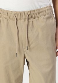 Pantalones de algodón beige con cordón ajustable y cintura elástica. Presentan una textura suave y un cordón beige en contraste con extremos de metal.