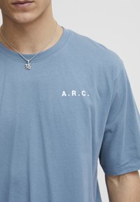 Hele sinine puuvillane t-särk ümara kaelusega, lühikeste varrukatega, mille esiküljel on valge tekst "A.R.C." ja hõbedane kaelakee sümboliga.