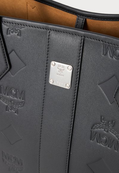 Sac en cuir noir avec logos MCM embossés et formes en losange, doté d'une plaque métallique argentée gravée MCM ainsi que d'un numéro de série.