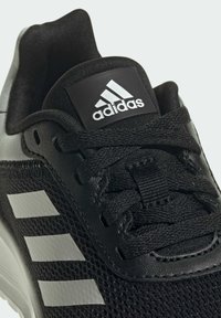 Scarpe sportive nere con una parte superiore in rete e sovrapposizioni sintetiche. Tre strisce bianche riflettenti sul lato, con una linguetta imbottita e lacci.