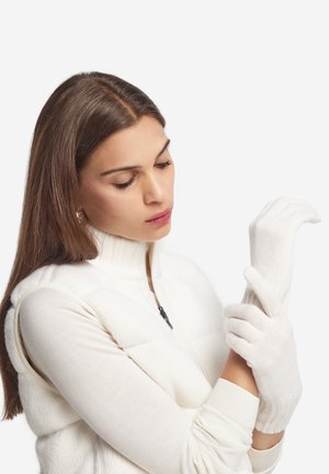 Des gants en tricot blancs avec des poignets côtelés et des ouvertures pour les doigts, portés par une personne dans un sweat à zip blanc, sur un fond uni.