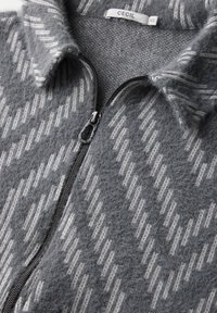 Veste en polaire grise zippée avec un motif texturé de lignes gris clair. Dispose d'un col doux et d'une fermeture éclair en métal à l'avant.