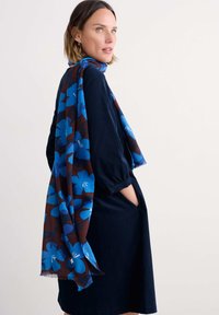 Ein blaues, floral gemustertes Tuch liegt über einem marineblauen Kleid, das eine weiche Textur, runde Ärmel und Seitentaschen aufweist. Das Tuch hat fransige Kanten.
