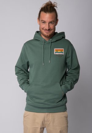 Green hoodie made of soft fabric, featuring a cassette tape design in vibrant colors on the chest pocket. Front kangaroo pocket included.