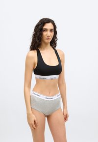 Γυναίκα με μακριά σγουρά μαλλιά, φοράει μαύρο αθλητικό σουτιέν και γκρι εσώρουχα Calvin Klein, στέκεται μπροστά σε μια απλή φωτεινή επιφάνεια.